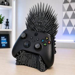 Pode incluir: Um comando sem fios Xbox preto pousado num suporte cinzento-escuro em forma de trono feito de numerosas espadas em miniatura. O comando possui um D-pad preto, dois joysticks anal&oacute;gicos e quatro bot&otilde;es de a&ccedil;&atilde;o coloridos A, B, X e Y.