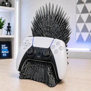 May include: A black, sword-themed controller stand shaped like a throne, featuring a high backrest of pointed blades. A white and black wireless game controller is resting securely in the center of the dark, textured, sword-covered base.