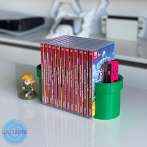 May include: A green, 3D-printed pipe-shaped game cartridge holder with a collection of Nintendo Switch game cases. The cases have red spines and game titles. A pink Joy-Con controller is visible. A small Link figurine is also present.