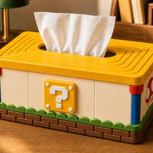 May include: A rectangular tissue box cover featuring a yellow top with concentric rectangular grooves. The sides are off-white with a yellow question mark block, blue corner pillars, a green scalloped base, and a brown brick-patterned bottom.