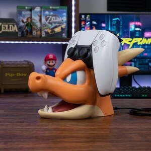 May include: An orange and cream-colored dragon-shaped controller stand featuring a wide-open mouth and horns, holding a white and black wireless game controller on its head. The stand has a smooth finish and is positioned on a dark wooden surface.