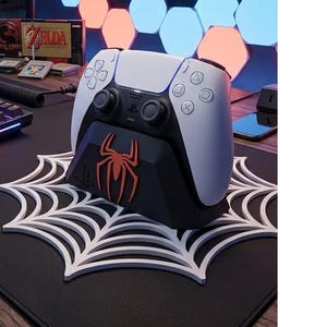 May include: A white and black wireless game controller sits on a black charging dock featuring a red spider emblem. The dock rests on a white, flat, circular spiderweb-shaped base. The controller has standard buttons, dual analog sticks, and a central PlayStation logo.