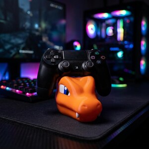 May include: An orange and white Charizard-themed controller stand supporting a black PlayStation controller. The stand is on a black surface, with a keyboard and computer in the background.