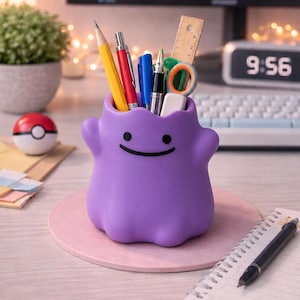 May include: A purple, blob-shaped desk organizer featuring a simple black dot-and-line face. The container holds various office supplies, including a yellow pencil, a red pen, a blue pen, a green pen, a wooden ruler, and a pair of small scissors.