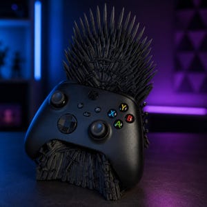 Pode incluir: Um comando sem fios Xbox preto pousado num suporte preto mate em forma de trono feito de numerosas espadas pontiagudas. O comando apresenta joysticks pretos, um pad direcional e bot&otilde;es de a&ccedil;&atilde;o A, B, X e Y multicoloridos.