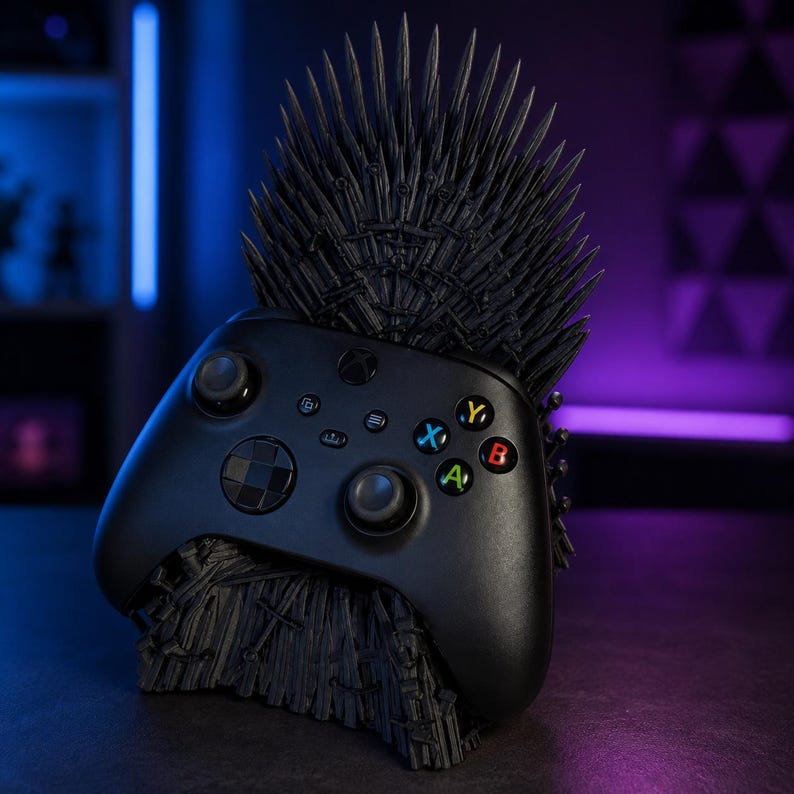 Pode incluir: Um comando sem fios Xbox preto pousado num suporte preto mate em forma de trono feito de numerosas espadas pontiagudas. O comando apresenta joysticks pretos, um pad direcional e bot&otilde;es de a&ccedil;&atilde;o A, B, X e Y multicoloridos.