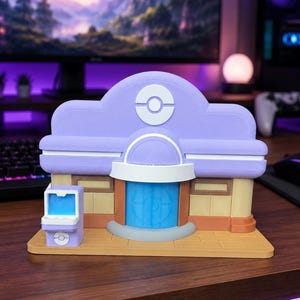 May include: A 3D printed model of a Pokemon Center building featuring a lavender roof with a white circular emblem, a blue central door, and tan walls. A small matching lavender kiosk with a blue screen sits to the left on a tan base.