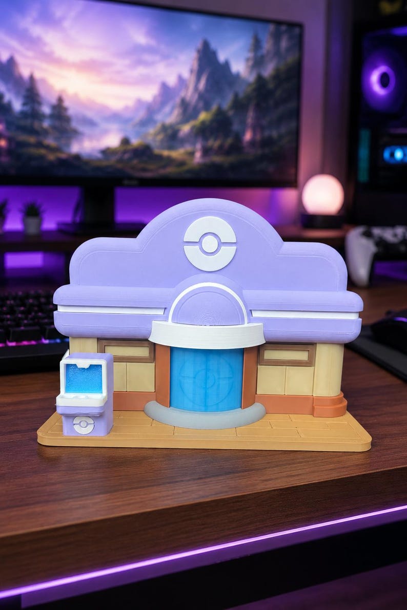 May include: A 3D printed model of a Pokemon Center building featuring a lavender roof with a white circular emblem, a blue central door, and tan walls. A small matching lavender kiosk with a blue screen sits to the left on a tan base.