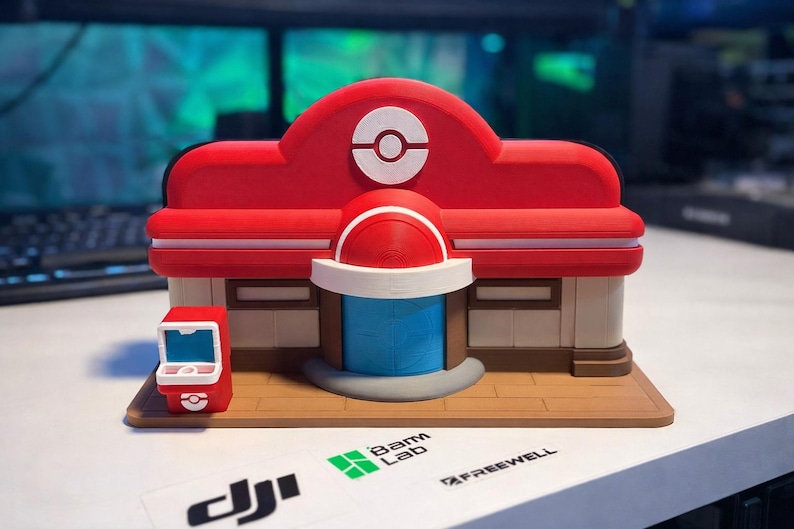 May include: A 3D-printed model of a red, white, and blue Pokemon Center building featuring a central white Poke Ball emblem above a blue door. A small red and white terminal stands to the left on a brown base. The structure has a smooth matte finish.