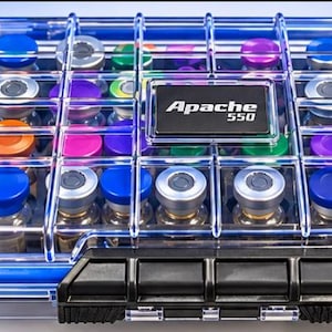 May include: A clear, rectangular Apache 550 protective case with a blue border and black latch, containing multiple small glass vials with colorful caps. The case features a grid-like structure and a black label with white text reading Apache 550.