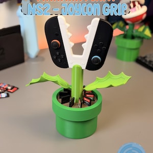 May include: A green Piranha Plant-themed Nintendo Switch Joy-Con grip. The grip is designed to hold the Joy-Cons, with the controllers forming the plant's jaws. The base is a green pot, and the design includes the text "Piranha Plant NS2 - Joycon Grip".
