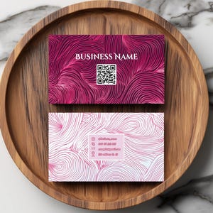 May include: Two business cards with a flowing, abstract design in shades of pink and white rest on a wooden tray. The top card features the text "BUSINESS NAME" and a QR code. The bottom card displays contact information.