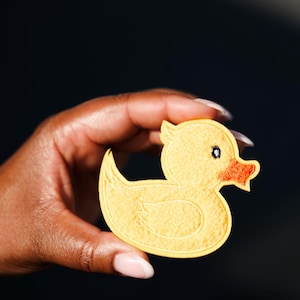 May include: A yellow felt duck-shaped applique with an orange beak and a small black eye. The applique is held in a hand against a dark background. The duck has a rounded body and a small tail.