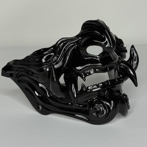 Puede incluir: Una máscara de samurái negra brillante con detalles intrincados. La máscara presenta dientes afilados, cuernos curvos y elementos similares al cabello. El diseño incluye aberturas para los ojos y la boca, y un remolino decorativo.