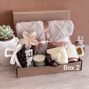 May include: A gift box containing self-care products. It features a folded beige blanket with a ribbon, a candle, a mug, soap, a flower, socks, and a succulent plant. The box is labelled "Box 2". The items are arranged in a brown cardboard box.