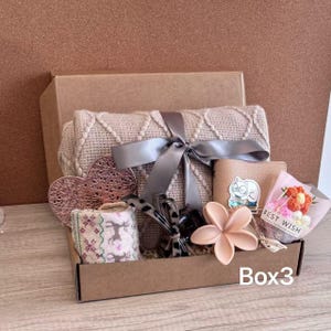 May include: A gift box filled with various items, including a beige blanket tied with a gray ribbon, a pink heart-shaped decoration, a wallet, a flower-shaped ornament, and a small bouquet. The box is labeled "Box3".
