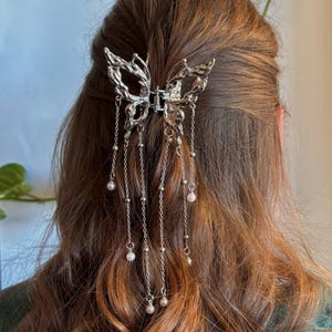 Peut inclure: Une pince à cheveux en métal argenté en forme de papillon avec une surface texturée rappelant du métal fondu. L'accessoire comporte plusieurs chaînes argentées pendantes de longueurs variées, ornées de petites perles blanches rondes.