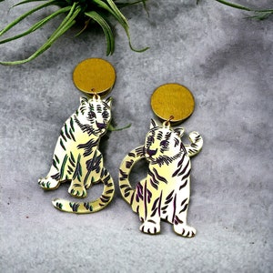 May include: A pair of gold-tone earrings featuring a white tiger design. The tigers are outlined in black and have purple accents. The earrings are attached to gold-tone circular posts.