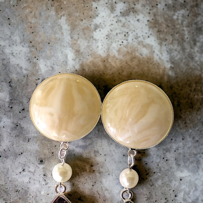 May include: A pair of cream-colored, round earrings with a silver trim. Each earring features a dangling pearl and a small, dark purple gemstone. The earrings are set against a gray, textured background.