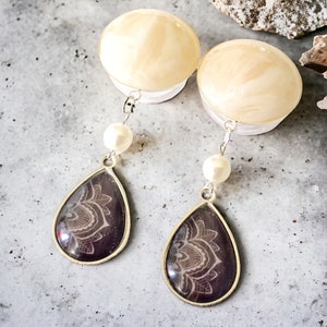 May include: A pair of dangle earrings featuring a teardrop-shaped pendant with a silver frame. The pendant has a dark purple background with a white floral design. Each earring has a white pearl and a cream-colored top.