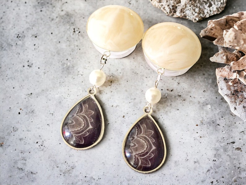 May include: A pair of dangle earrings featuring a teardrop-shaped pendant with a silver frame. The pendant has a dark purple background with a white floral design. Each earring has a white pearl and a cream-colored top.