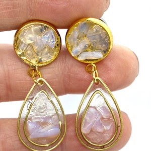 Peut inclure: Boucles d'oreilles en forme de goutte d'eau dorées. Les boucles d'oreilles sont en résine transparente avec des éclats irisés incrustés. Le haut des boucles d'oreilles est rond avec un design similaire.
