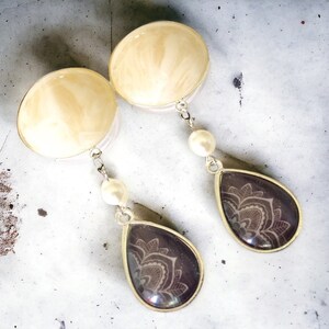 May include: A pair of dangle earrings. Each earring features a large, round, cream-colored top with a silver-colored border. A silver chain connects to a teardrop-shaped pendant with a dark purple background and a white floral design. A white pearl is attached to the chain.