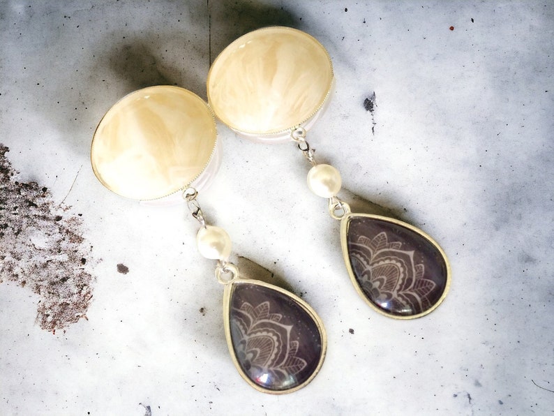 May include: A pair of dangle earrings. Each earring features a large, round, cream-colored top with a silver-colored border. A silver chain connects to a teardrop-shaped pendant with a dark purple background and a white floral design. A white pearl is attached to the chain.