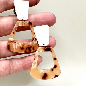 May include: A pair of dangle earrings with a brown and white tortoiseshell pattern. The earrings have a white triangle-shaped piece at the top and a larger, open, triangle-shaped piece below.