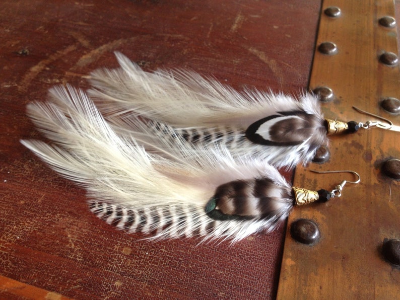 May include: A pair of feather earrings with black and white feathers and gold accents. The earrings are on a wooden surface.