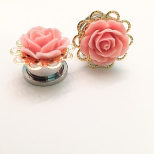 K&ouml;nnte beinhalten: Zwei Ohrstecker in Form einer rosa Rose mit goldener Filigranverzierung. Die Stecker sind aus Edelstahl und haben eine gl&auml;nzende Oberfl&auml;che.