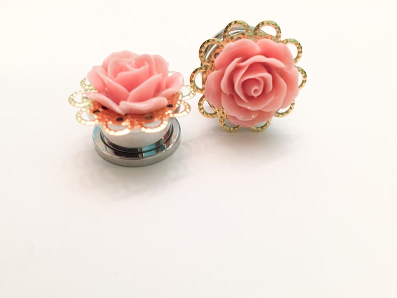 K&ouml;nnte beinhalten: Zwei Ohrstecker in Form einer rosa Rose mit goldener Filigranverzierung. Die Stecker sind aus Edelstahl und haben eine gl&auml;nzende Oberfl&auml;che.