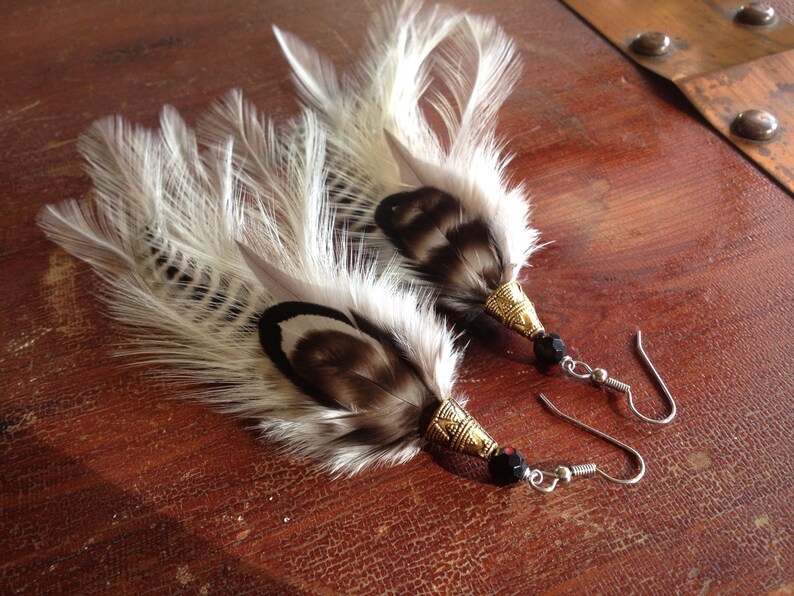 May include: A pair of feather earrings with black beads and gold accents. The feathers are white with black markings.