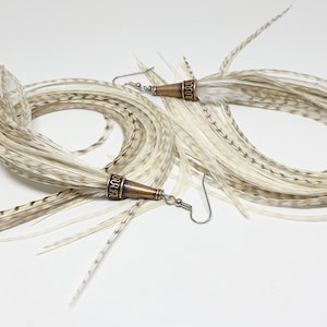 Puede incluir: Un par de pendientes largos de plumas blancas con detalles marrones. Los pendientes tienen una cuenta de cobre de forma cónica en la parte superior con un diseño decorativo.