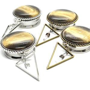 May include: A pair of brown and white striped ear gauges with silver and gold triangle charms. The charms have a clear crystal hanging from them.