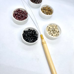 May include: An assortment of hair extension beads in various colors, including black, brown, and blonde, displayed in small white containers. A wooden-handled hair extension tool is also present, designed for applying the beads to hair.