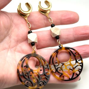 May include: A pair of gold ear cuffs with a dangling chain and a brown and black tortoiseshell patterned circle pendant. The chain features black and white beads.