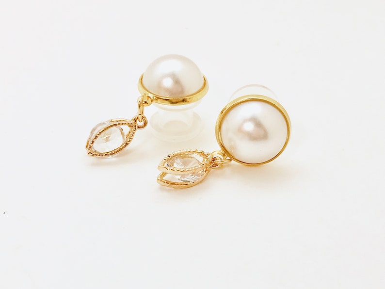 May include: A pair of gold earrings with a pearl and a clear crystal drop. The earrings are on a white background.