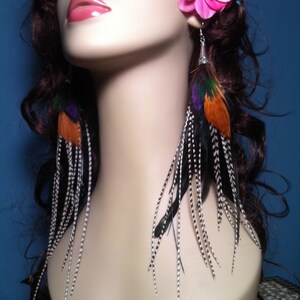 Puede incluir: Un par de pendientes largos de plumas con plumas negras, blancas, naranjas y moradas. Los pendientes cuelgan de un gancho plateado.