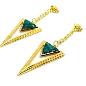 May include: A pair of gold-colored earrings with a geometric design. Each earring features a gold triangle with a smaller triangle filled with teal-colored, textured material. The earrings hang from a delicate gold chain.