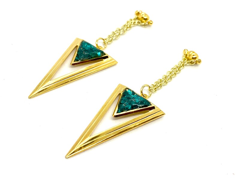 May include: A pair of gold-colored earrings with a geometric design. Each earring features a gold triangle with a smaller triangle filled with teal-colored, textured material. The earrings hang from a delicate gold chain.