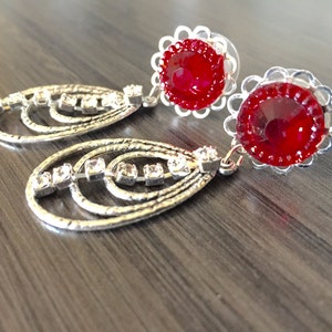 Puede incluir: Un par de pendientes colgantes con un strass rojo y un diseño de filigrana plateada. Los pendientes tienen una forma ovalada plateada con una fila de strass transparentes.