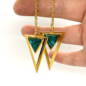 May include: Gold-tone earrings featuring a double triangle design. The outer triangle is a solid gold color, while the inner triangle is filled with a sparkling teal material. The earrings hang from a delicate gold chain.