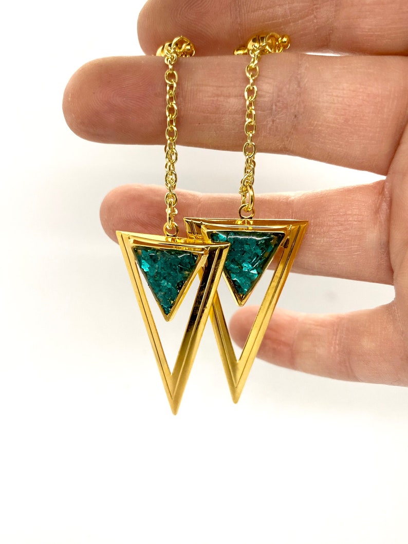 May include: Gold-tone earrings featuring a double triangle design. The outer triangle is a solid gold color, while the inner triangle is filled with a sparkling teal material. The earrings hang from a delicate gold chain.