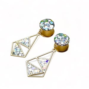 May include: A pair of gold ear gauges with a geometric design and iridescent glitter accents. The gauges are round and have a dangling geometric design.