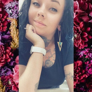 May include: Gold-tone triangle earrings with a turquoise inlay, hanging from delicate chains. The earrings are worn by a person wearing a black top and a white watch. The background features a floral arrangement with deep red and purple flowers.