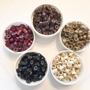 May include: Five white bowls filled with hair extension rings in various colours: burgundy, dark brown, light brown, black, and blonde. The rings are small, cylindrical, and have a silicone lining.