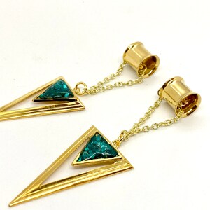 May include: A pair of gold-colored ear gauges with a geometric design. Each gauge features a triangular pendant with a teal-colored inlay, connected to a gold chain and a cylindrical end. The gauges are displayed against a white background.