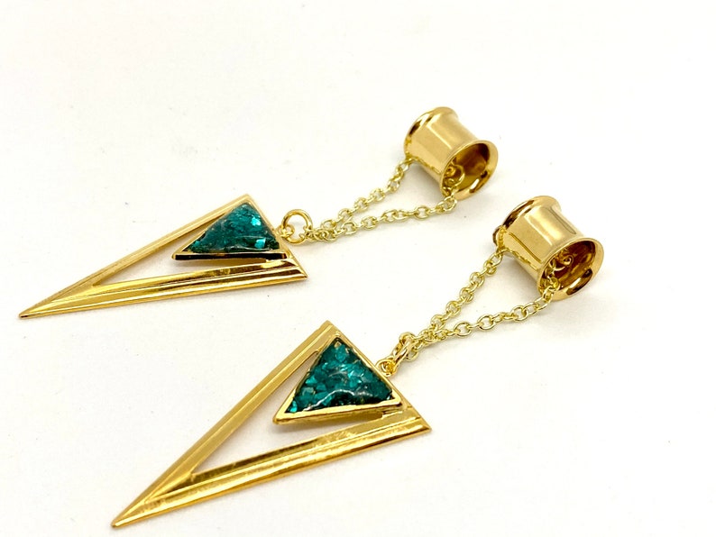 May include: A pair of gold-colored ear gauges with a geometric design. Each gauge features a triangular pendant with a teal-colored inlay, connected to a gold chain and a cylindrical end. The gauges are displayed against a white background.