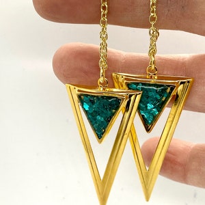 May include: Gold-tone earrings featuring open triangle pendants filled with sparkling teal-colored material. The earrings hang from delicate gold chains. The design is geometric and modern, suitable for everyday wear or special occasions.
